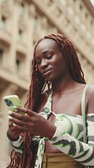 Wall Mural - VERTICAL VIDEO: Smiling gorgeous woman with African braids wearing top stands on the sidewalk next to the road and uses mobile phone. Stylish girl writes message on smartphone