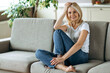 © Kateryna - Smiling caucasian middle aged blonde woman sits on sofa at living room dressed in casual wear looks at camera and smiling, rest from homework, weekend