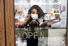Curly Female African American Small Business Owner In Medical Mask Points At A Signboard WE ARE OPEN At The Entrance To A Restaurant, Bar, Cafe Or Shop, And Waiting For Visitors