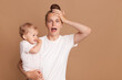 Indoor shot of young tired shocked mother holding a child in her arms, female wearing white casual style T-shirt posing with a baby isolated on brown background, screaming, keeps hand on head.