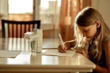 A 6-7-year-old Girl With Blonde Hair In A Green T-shirt Is Sitting At Home At A Table And Drawing With Markers. The Girl Is Engaged In Creativity. She's Learning To Draw
