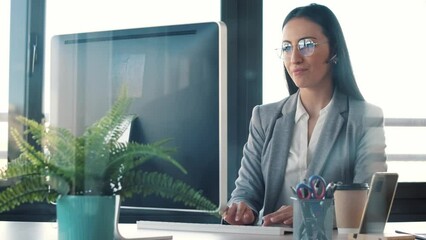 Wall Mural - Video of beautiful business woman making video call with computer while talking with earphone sitting in modern startup office.