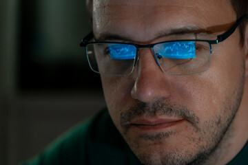 Portrait of millennial concentrated trader man in glasses looks at screen with graphs in dark. Caucasian man monitors exchange rate. Business Graph and chart of currency reflection in eyeglasses.