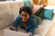 © Wavebreak Media - Smiling young african american woman using laptop while lying on sofa at home, copy space