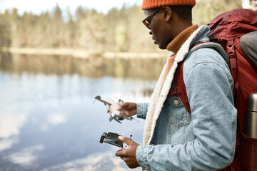 Young african american man tourist holding control console for quadcopter in hands, filming nature hiking in wild forest by the river or lake, carrying backpack on shoulders. Traveling concept