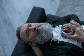 Wall Mural - top view of disappointed senior man with beard lying on couch with glass of whiskey while suffering crisis.