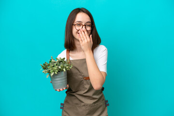 Wall Mural - Gardener Ukrainian woman holding a plant isolated on blue background happy and smiling covering mouth with hand