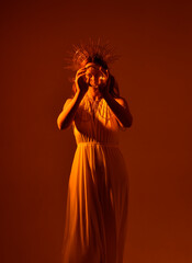 Close up portrait of beautiful red-haired woman wearing long flowing fantasy toga gown with golden halo crown jewellery,  creative hand gestures on a dark moody background with glowing orange lighting
