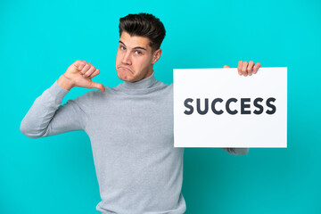 Young handsome caucasian man isolated on blue bakcground holding a placard with text SUCCESS with proud gesture