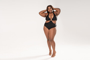 Full-length photo of beautiful plus size dark skinned woman posing on light gray, wearing black fashionable swimsuit.