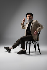 Poster - full length of elegant senior man in beige jacket and derby hat sitting on wooden chair on grey.