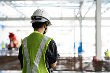 An Asian female engineer wearing a reflective green safety vest wears a protective hard hat. Standing supervisor at a construction site with construction workers working in the background.