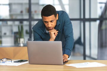 Concentrated Afro man working using laptop in modern office. Successful business African American man, looking at computer screen, analyzing something