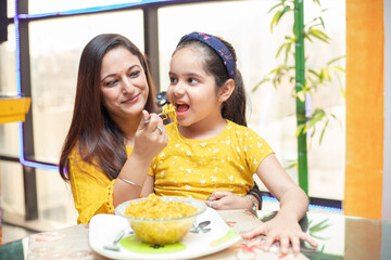 young indian mother feed her little daughter noodles breakfast at table, little girl child having fo
