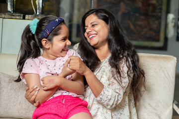 happy playful indian mother and daughter sitting together at home, love and care, asian woman with g
