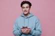 © Tatiana - a handsome, attractive man stands on a pink background in a light blue hoodie and holding a smartphone in his hands looks at the camera with a friendly smile