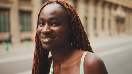 Wall Mural - Close-up of beautiful woman with African braids raising her head and looking at the camera with smile on the building background.