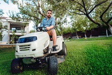 Gardener Driving A Riding Lawn Mower In A Garden. Professional Landscaper Using Tractor At Mowing Lawn.