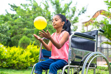 Concept Of Happiness, Freedom And Enjoyment Showing By Smiling Alone Girl Kid With Disability Playing With Ball While On Wheelchair At Park.
