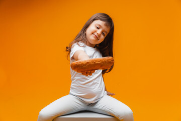 Wall Mural - a little girl sits on a cube on a yellow background and eats a loaf. healthy food, natural products. bread