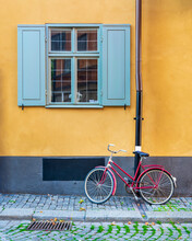 Red Bicycle Parked Under Old Vintage Traditional Window With Blue Shutters In An Orange Wall At A Narrow Cobblestone Street In The Old Town Of Gamla Stan, Stockholm, Sweden