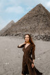 Young redhead tourist girl in brawn dress with a leather backpack standing on the sand in Egypt, Giza. Pyramids of Khafre and Cheops on backround