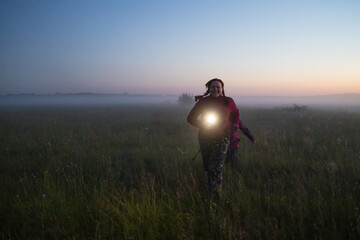 Attractive body positive woman and child run through the meadow at sunset, shining a flashlight. Happy summer evening.
