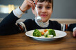 © Taras Grebinets - Focus on steamed vegetables, healthy vegan food against a blurred adorable boy holding fork and smiling looking at camera during lunch in cafeteria