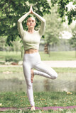 Fototapeta Las - A young beautiful slender girl with long blonde hair does yoga in the summer in nature by the pond in the park.