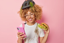 Happy Curly Haired Active Woman Cyclist Rests After Riding Bike Scrolls Newsfeed Via Smartphone Eats Tasty Hamburger Dressed In Dirty Clothes Isolated Over Pink Background. Fast Food Concept