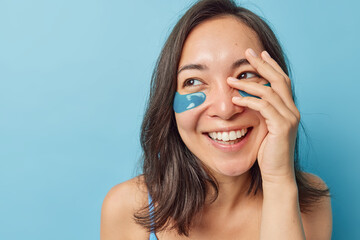 Wall Mural - Happy brunette eastern woman applies beauty patches to reduce fine lines and wrinkles makes face palm smiles joyfully looks away poses against blue background with copy space area for your text