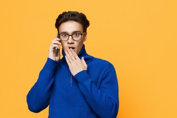 Wall Mural - horizontal photo of a cute, surprised man standing on an orange background in a blue zip-up jacket and black eyeglasses, talking on the phone with his hand covering his mouth.