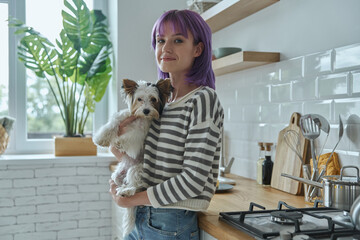 Wall Mural - Beautiful young woman carrying her cute little dog while standing at the kitchen