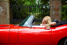 Beautiful Blonde Lady Leaning On The Door Of A Red  E-type Jaguar Looking At Herself In The Wing Mirror