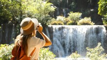 Female Travel Blogger With Photo Camera Shooting Amazing Tropical Waterfall In Asia Journey. Cinematic And Inspiration Video Of Photographer Woman Taking Picture Of Nature In Travel Vacation.