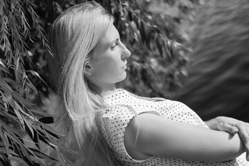 Wall Mural - Black and white portrait a young beautiful blonde Caucasian girl in a polka-dot dress against the background of a river and a willow branch, looking down. Hard sunlight, day. Nature. Summer