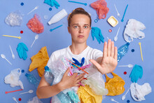 No Plastic And Environmental Pollution. Serious Caucasian Woman Pulls Palm Forward, Wearing White T-shirt, Asking Not To Pollute Our Planet, Posing With Garbage In Hands Against Blue Wall.