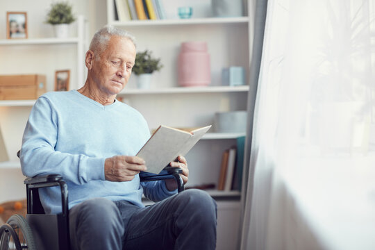 Serious handsome senior man in light blue sweater sitting in wheelchair and reading book in living room
