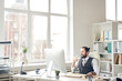 © Mediaphotos - Cheerful confident young hipster businessman with ponytail sitting at table and drinking water while working with computer