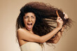 © SHOTPRIME STUDIO - HAIR LOSS. TANGLED HAIR CONCEPT. Screaming suffering irritated awesome curly Latin lady with hairbrush combing hair posing isolated on pastel beige background, look aside. Copy space, cool offer