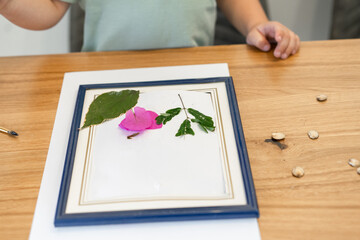 Wall Mural - Child with white paper, picture frame, leaves, and flowers making wildflowers greeting card on dining wood table in home.