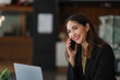 © ArLawKa - Successful young Asian businesswoman holding a smartphone working on a laptop computer financial graph data on the desk in the office.