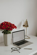 Laptop computer with blank screen on table with glasses, lamp, red gerber flowers bouquet. Aesthetic influencer styled workspace interior design template with mockup copy space