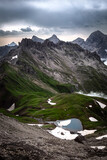 Fototapeta  - Mountain Valley with green meadows, a blue lake and snow in dramatic light in the mountains of the alps in Allgäu, Germany