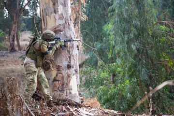 The military hid behind a tree and fired at the enemy. A guy in camouflage is hiding behind a tree against the backdrop of a cliff. Airsoft game in nature