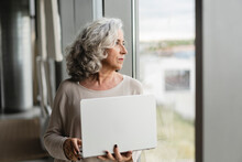 Senior Businesswoman With Laptop Looking Out Of Window At Office