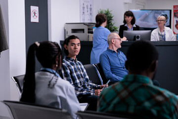 Portrait of asian patient waiting for doctor appointment in private hospital while doctor is talking with diverse people. Young adult in medical clinic attending doctor appointmet for checkup.