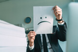 Fototapeta  - Unhappy businessman holding paper with frowning emoticon in office interior