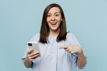 Wall Mural - Young happy shocked fun woman she 20s in casual blouse hold in hand use point index finge on mobile cell phone isolated on pastel plain light blue background studio portrait. People lifestyle concept.