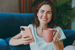 © ViDi Studio - Young happy pensive fun woman she 20s in casual clothes mint t-shirt use mobile cell phone hold cup drink coffee tea in morning look camera sit on blue sofa indoor rest at home in own room apartment
